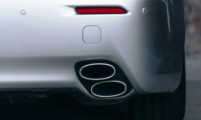 Doppio scarico cromato su un'auto sportiva argentata, parte del collettore di scarico. Sensori di parcheggio e fanale posteriore rosso visibili.