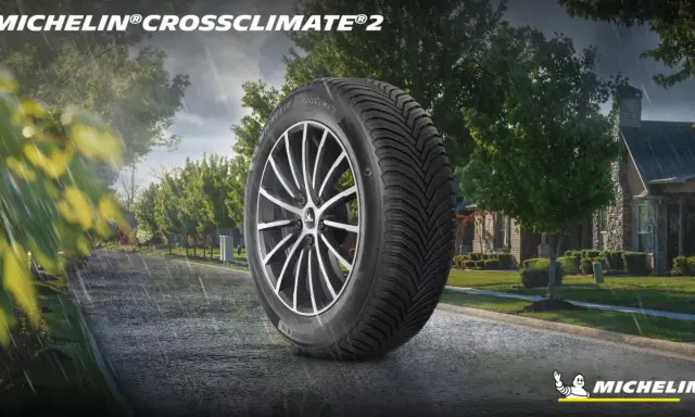 Pneumatico Michelin CrossClimate 2 in primo piano su strada bagnata, con case e alberi sullo sfondo. Nessun difetto visibile.