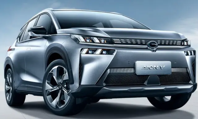 Un SUV grigio metallizzato, l'Aion V, con un design moderno e fari a LED affilati, si presenta su uno sfondo blu cielo.