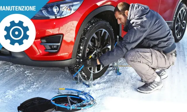 Uomo monta catene da neve su una ruota di un'auto rossa in un paesaggio innevato. Le gomme invernali sono essenziali per la sicurezza.