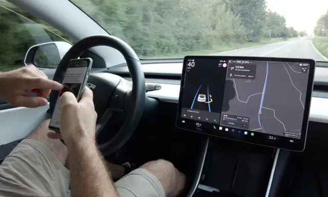 Mani sul volante, ma lo sguardo è sullo smartphone mentre il **Tesla Autopilot** guida. Navigazione e musica sul grande schermo.