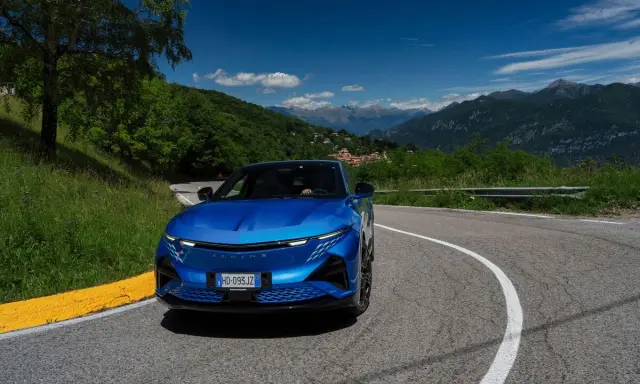 L'Alpine A290 sfreccia su una strada di montagna panoramica, con un cielo azzurro e montagne sullo sfondo.