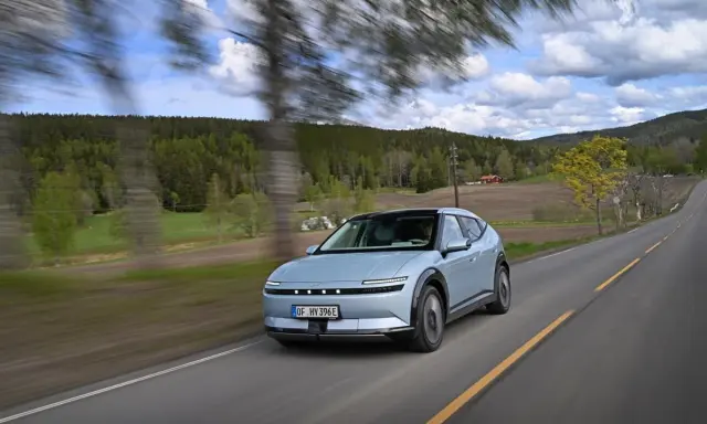 La Hyundai Ioniq 5 sfreccia su una strada panoramica tra alberi e colline verdi, con un cielo nuvoloso.