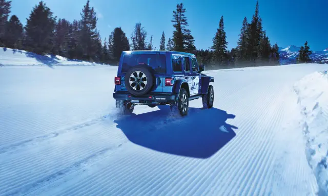 Un blu Jeep Wrangler affronta la neve con pneumatici Toyo Open Country AT3, lasciando tracce su un sentiero battuto tra alberi innevati e montagne.