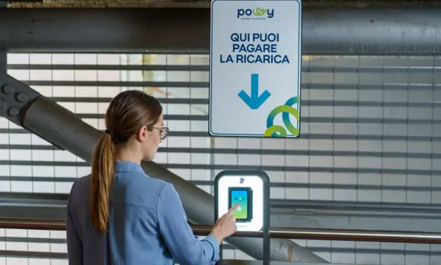 Donna interagisce con un terminale per pagare la ricarica dell'auto elettrica. Il cartello indica "Qui puoi pagare la ricarica", suggerendo il costo ricarica auto elettrica.