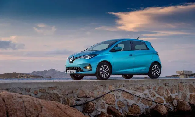 Un'elegante Renault Zoe blu, uno dei tanti modelli Renault, è parcheggiata su un muretto di pietra con il mare e le montagne sullo sfondo.