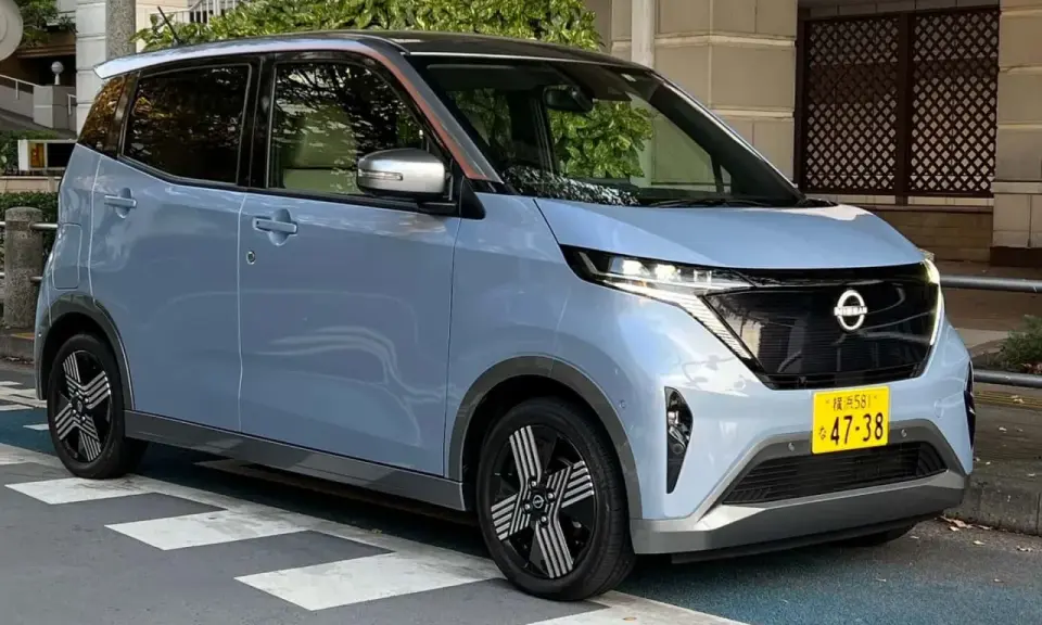 Nissan Sakura azzurro pastello, un'auto elettrica compatta con dettagli color rame sul tetto, parcheggiata su strisce pedonali.