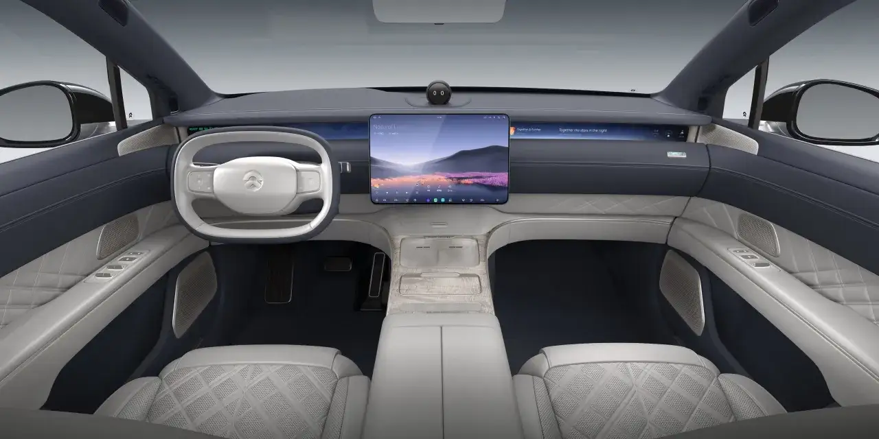Interno lussuoso del NIO ET9 con sedili in pelle trapuntata, volante bianco e ampio display centrale che mostra un paesaggio sereno.
