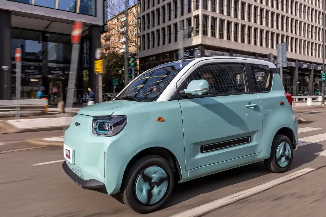 Piccola auto elettrica 4 posti color menta, in movimento su strada urbana con edifici moderni sullo sfondo.