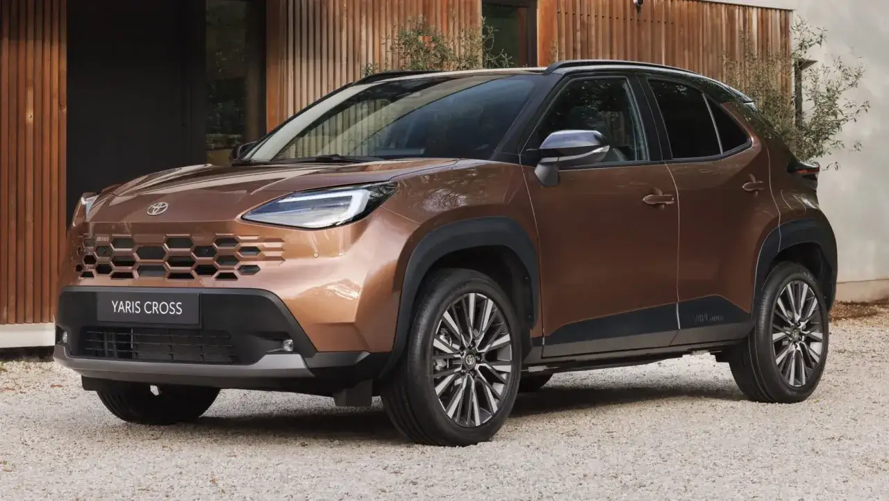 Nuova Yaris Cross 2026 color bronzo, con griglia anteriore a nido d'ape e cerchi in lega bicolore, parcheggiata su ghiaia davanti a una casa moderna.