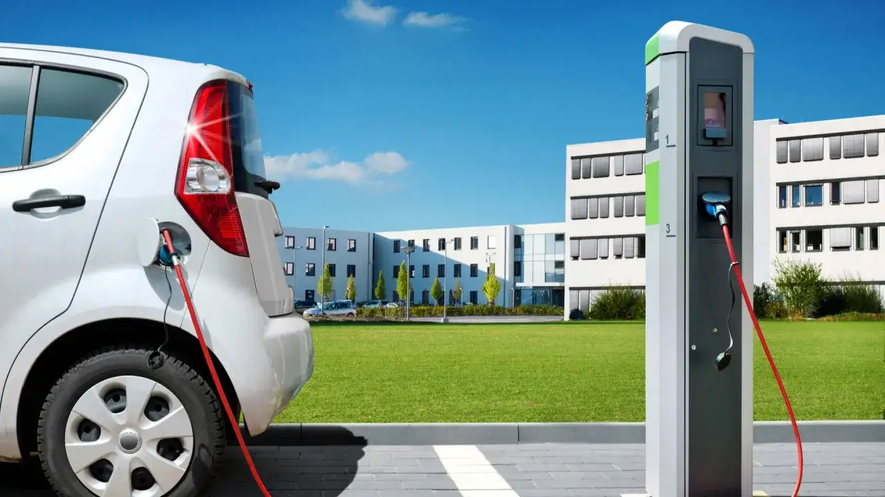 Un veicolo elettrico bianco è collegato a una colonnina di ricarica. Sullo sfondo, un moderno edificio con un prato verde.