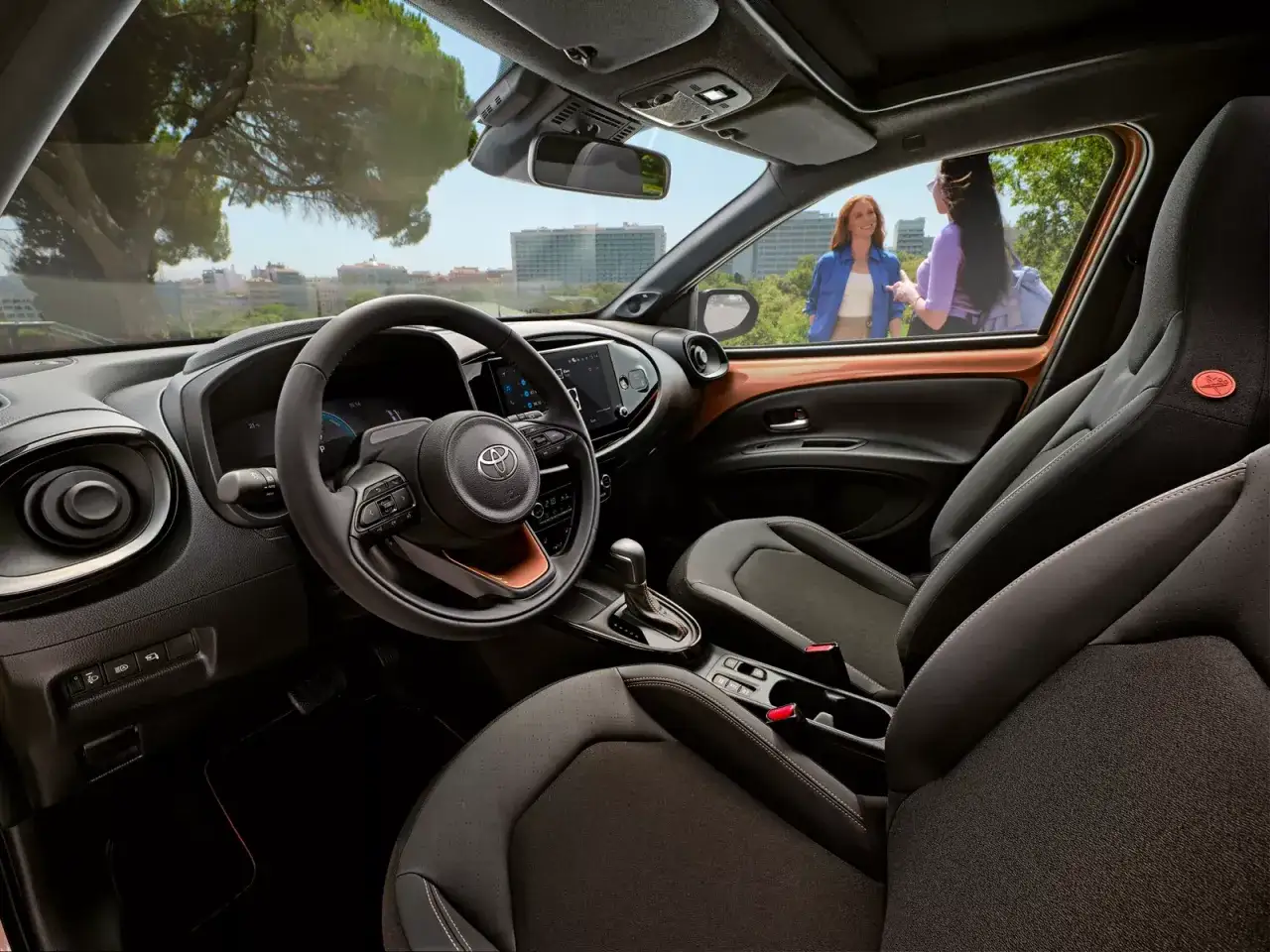 Interni moderni della Toyota Aygo X con sedili sportivi e dettagli color rame. Due donne chiacchierano all'esterno.