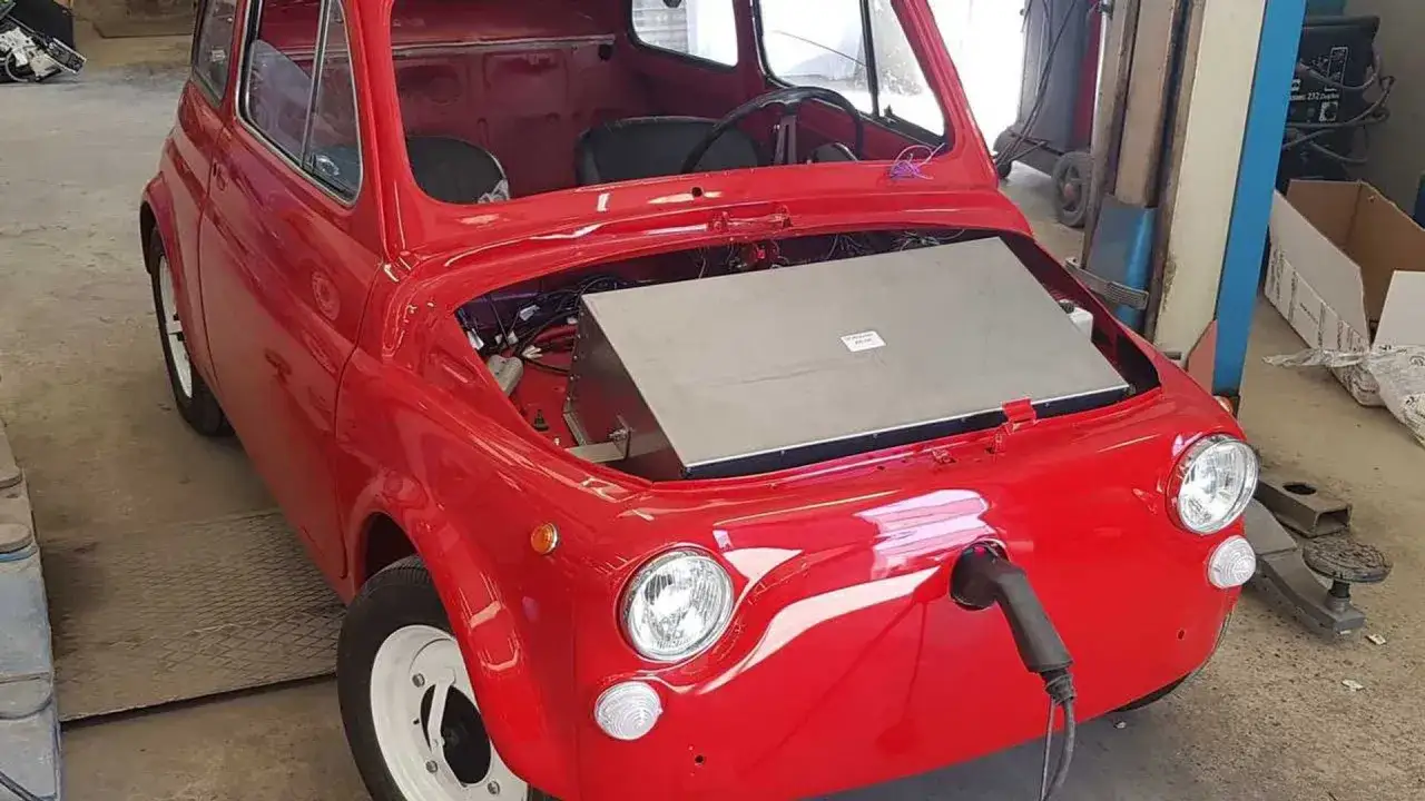 Un'iconica Fiat 500 rossa in fase di trasformazione auto in elettrica, con un pacco batterie visibile nel vano motore e un cavo di ricarica collegato.