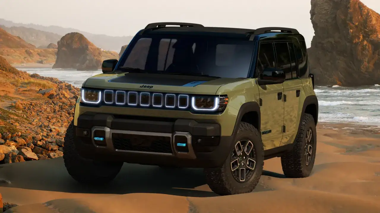 Un fuoristrada elettrico Jeep color kaki si ferma sulla sabbia dorata, con onde che lambiscono la costa rocciosa sullo sfondo.