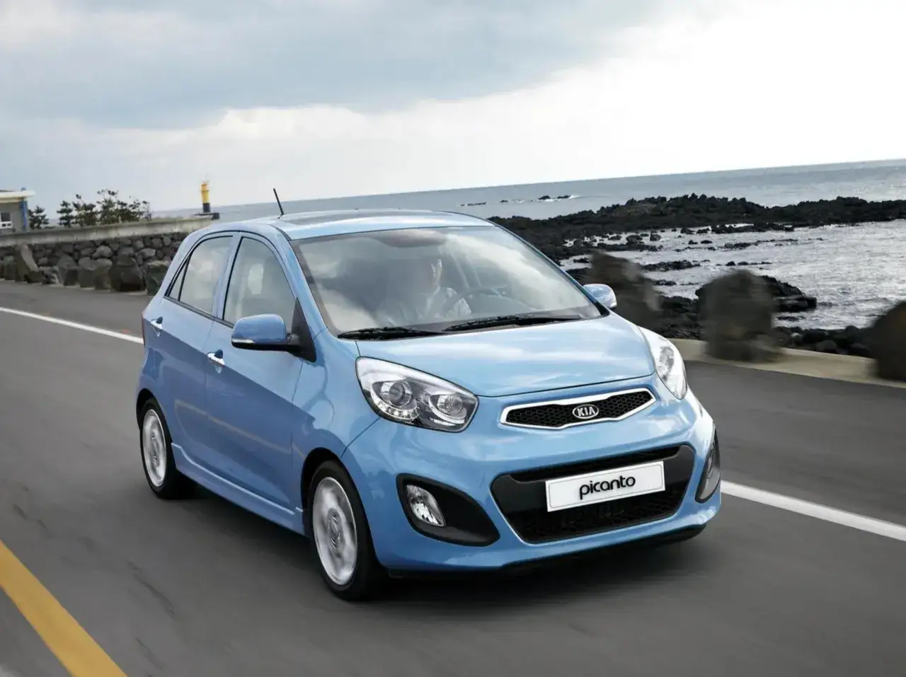La Kia Picanto blu sfreccia lungo una strada costiera. Nonostante i suoi piccoli difetti, &egrave; un'auto agile e divertente da guidare.