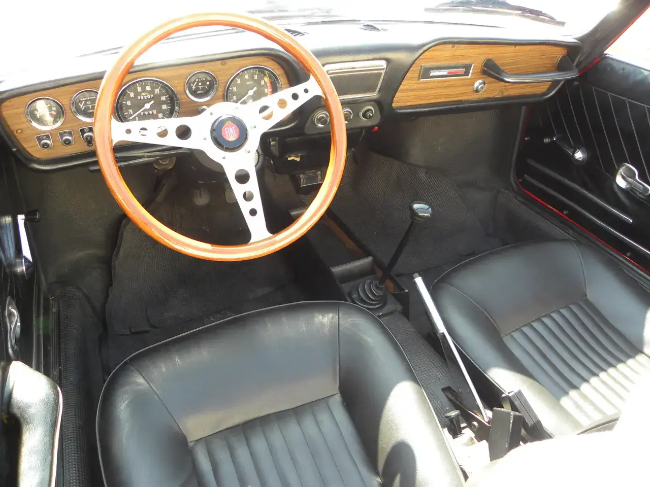 Interno auto d'epoca con volante in legno, cruscotto con indicatori e leva del cambio. Il freno a mano ghiacciato è visibile tra i sedili in pelle nera.