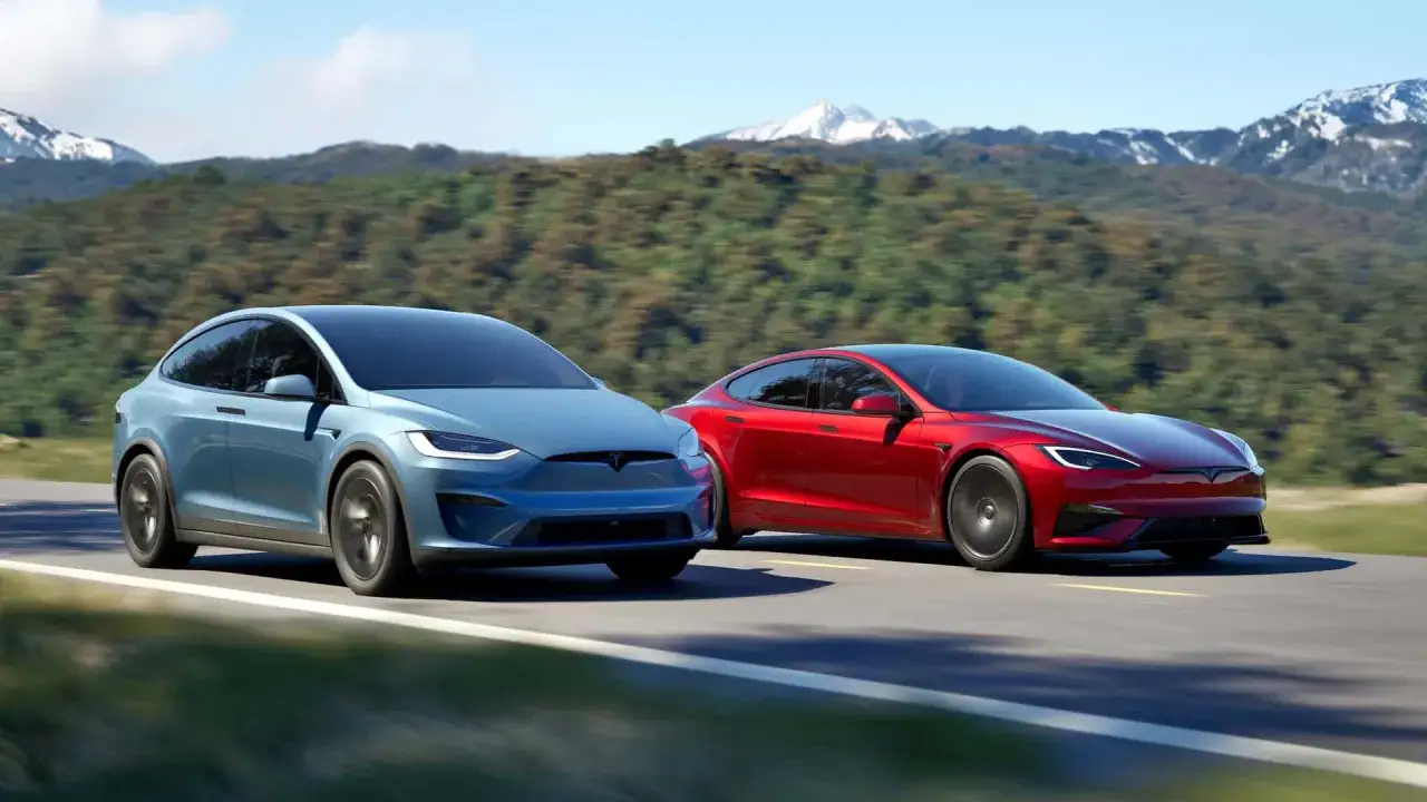 Due Tesla, una blu (SUV) e una rossa (berlina), sfrecciano su una strada panoramica. La berlina rossa, la Tesla più costosa, spicca per il suo design sportivo.