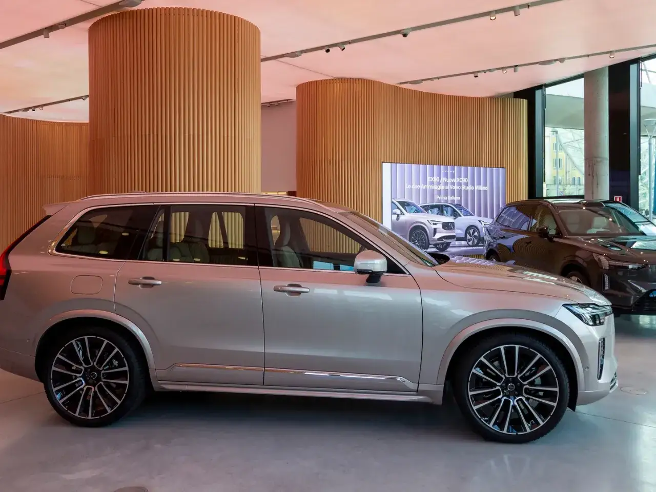 Volvo XC90 color argento, in esposizione in uno showroom moderno con colonne di legno e un grande schermo che mostra altri modelli.