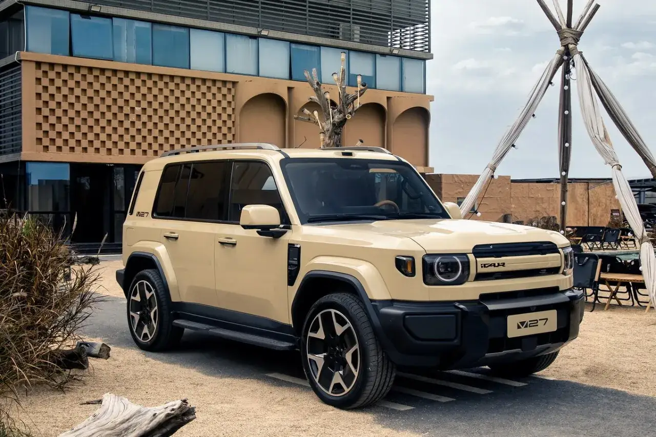 Jeep cinese color sabbia, modello V27, parcheggiata davanti a un edificio moderno con elementi decorativi in mattoni e un teepee decorato con luci.