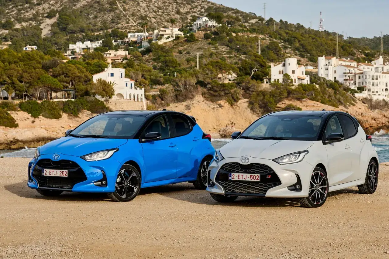 Due Toyota Yaris, una blu e una grigia, con diverse motorizzazioni, parcheggiate su una spiaggia con case bianche sullo sfondo.