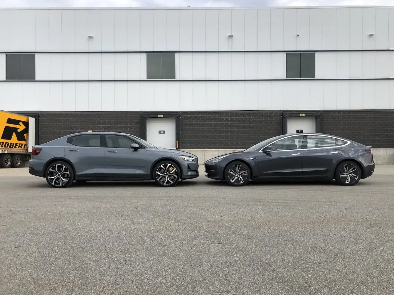 Due auto elettriche grigie, una Polestar 2 e una Tesla Model 3, parcheggiate davanti a un magazzino. Si attende l'uscita della Tesla Model 2.