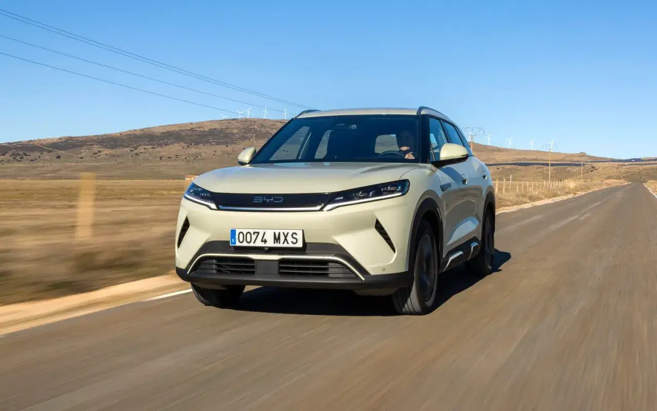 Un SUV BYD color crema sfreccia su una strada desolata, con pale eoliche sullo sfondo.