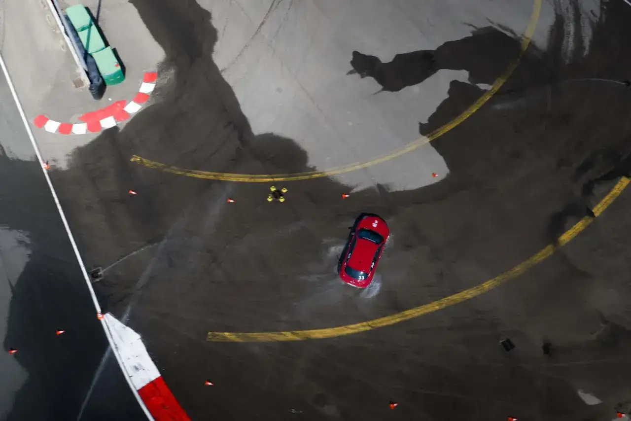 Auto rossa in derapata su asfalto bagnato, dimostrando le migliori gomme su bagnato. Coni arancioni e segnaletica a terra.