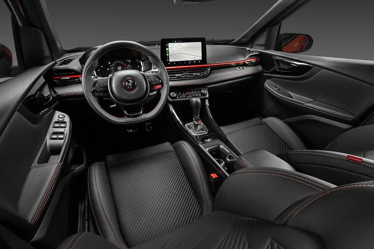 Interni sportivi della Fiat Fastback 2026: sedili in pelle nera con cuciture rosse, volante multifunzione e ampio display centrale.