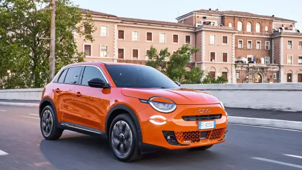Auto elettrica arancione in movimento, con 400 km di autonomia, su strada urbana con edifici storici sullo sfondo.